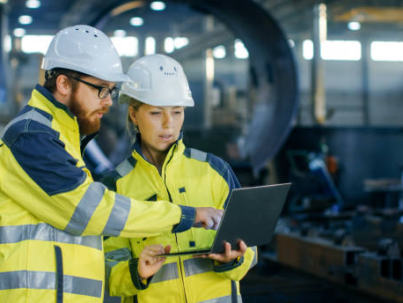 Male and Female Industrial Engineers in Hard Hats Discuss New Project while Using Laptop. They Make Showing Gestures.They Work in a Heavy Industry Manufacturing Factory.