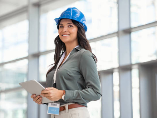 Beautiful mid adult woman architect wearing blue hardhat at construction site while working on digital tablet. Supervisor wearing safety helmet while working in a building site. Successful and proud inspector looking away with copy space.