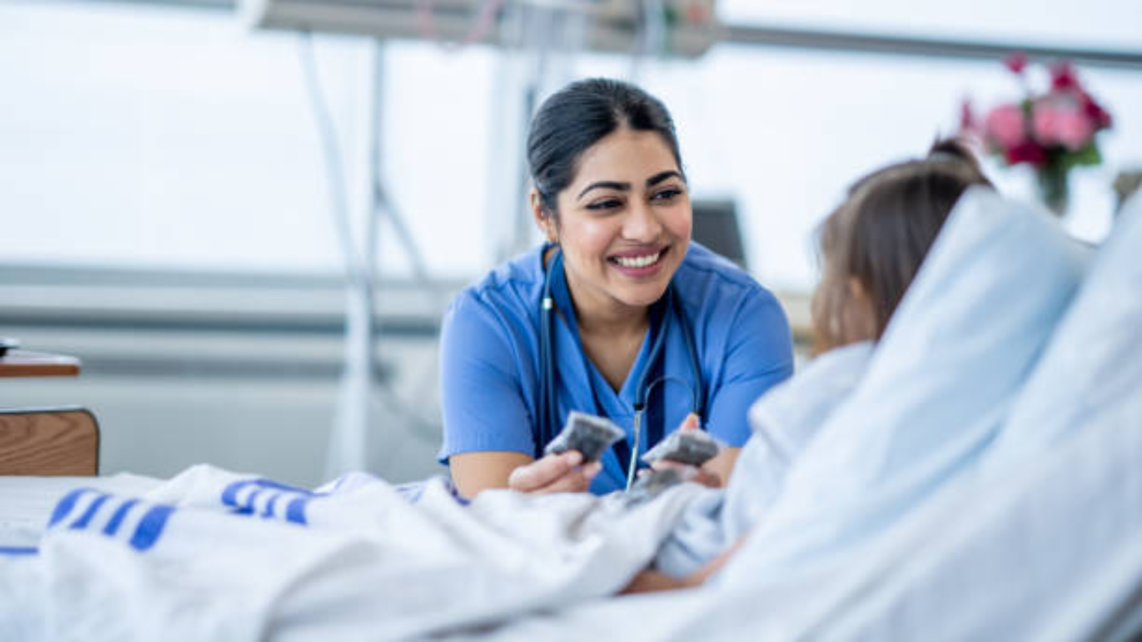 A female nurse of Middle Eastern decent sits at the edge of a hospital bed as she check in on her young patient. She is wearing blue scrubs and is attempting to cheer the young girl up.