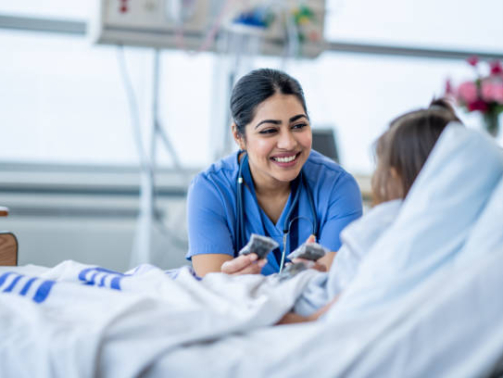 A female nurse of Middle Eastern decent sits at the edge of a hospital bed as she check in on her young patient. She is wearing blue scrubs and is attempting to cheer the young girl up.